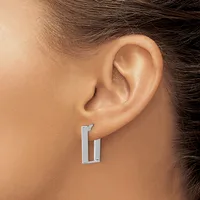 A person wearing a pair of rectangular hoop earrings in polished silver metal, shown from the side view near the ear.
