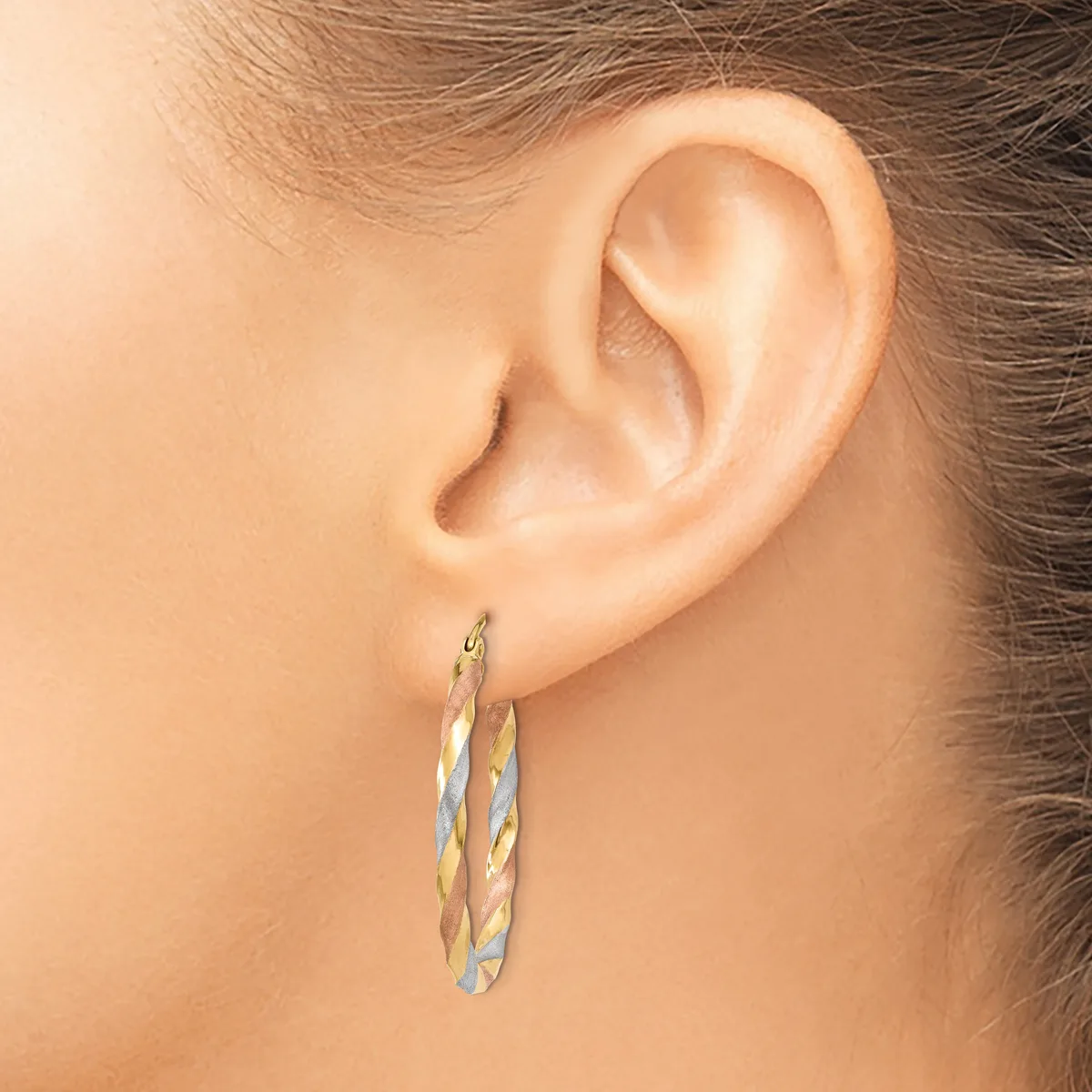 A woman's ear wearing a pair of twisted oval hoop earrings in two-tone gold and silver metal.