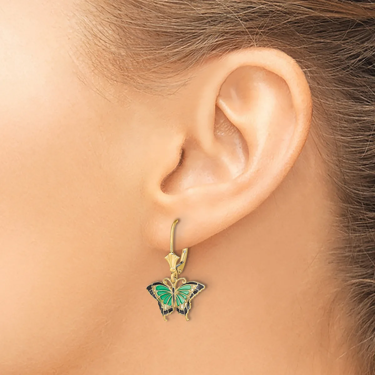 A person wearing a gold hook earring with a green and black enameled butterfly pendant hanging from their earlobe.