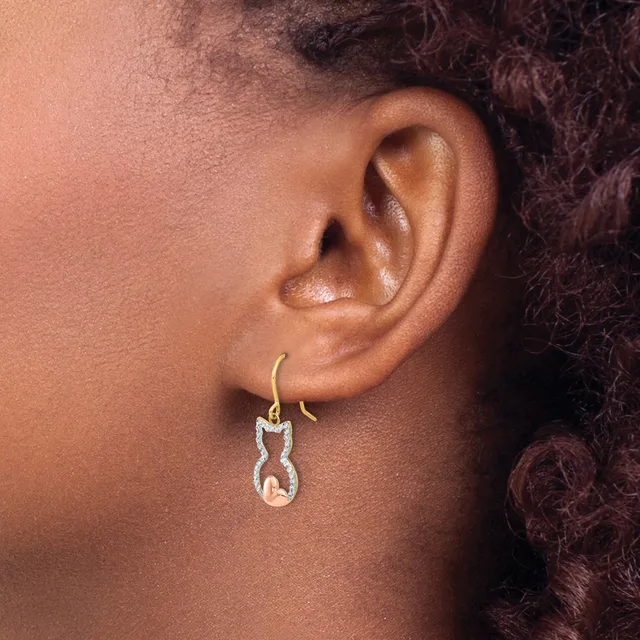 A person wearing a gold hook earring with a white diamond-outlined cat pendant hanging from it.
