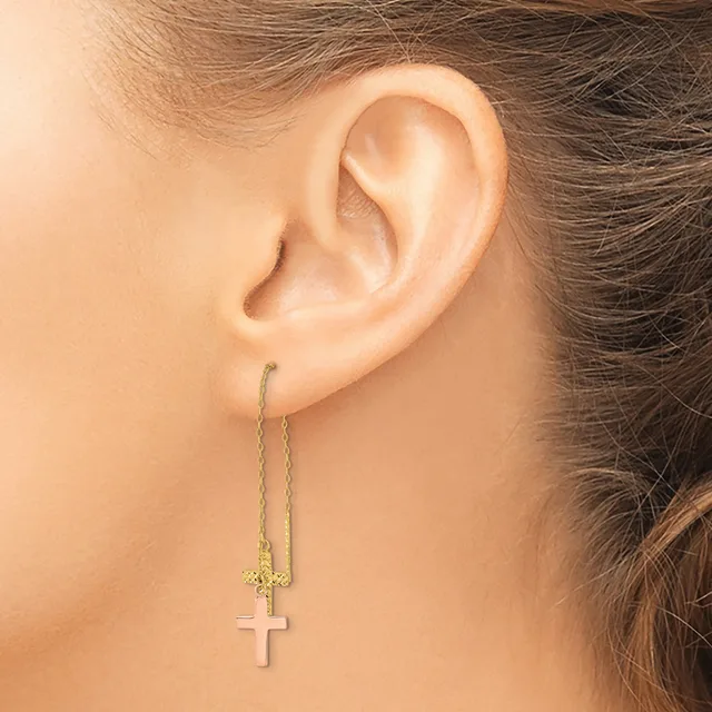 A person wearing a long chain earring with a small cross pendant in rose gold, shown from the side profile near their ear.