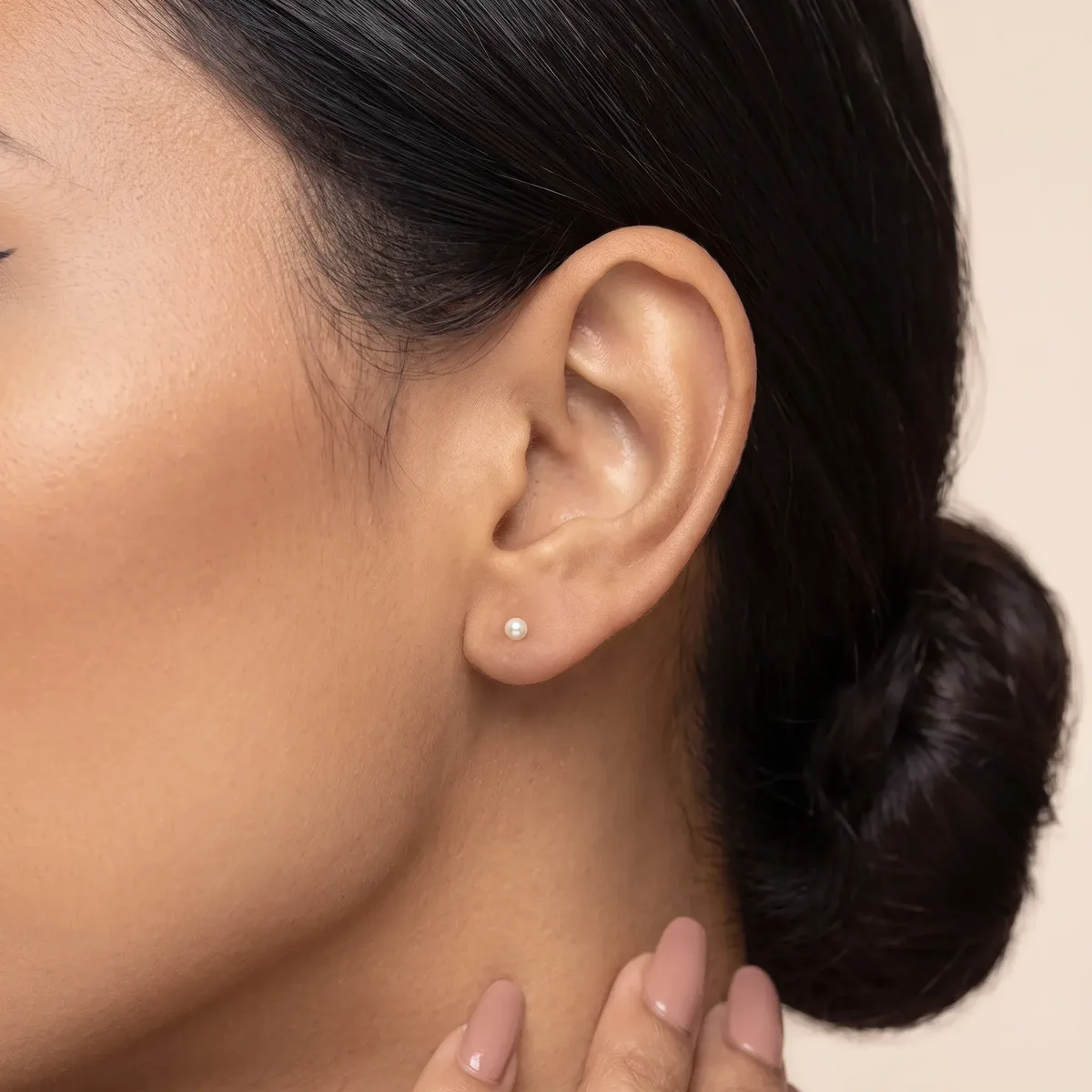 A person with dark hair in a bun wears a small pearl stud earring in their earlobe, shown in profile against a neutral background.