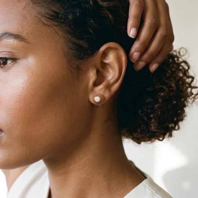 A person with curly brown hair pulled back wears a simple pearl stud earring visible in their earlobe, shown in profile against a white background.