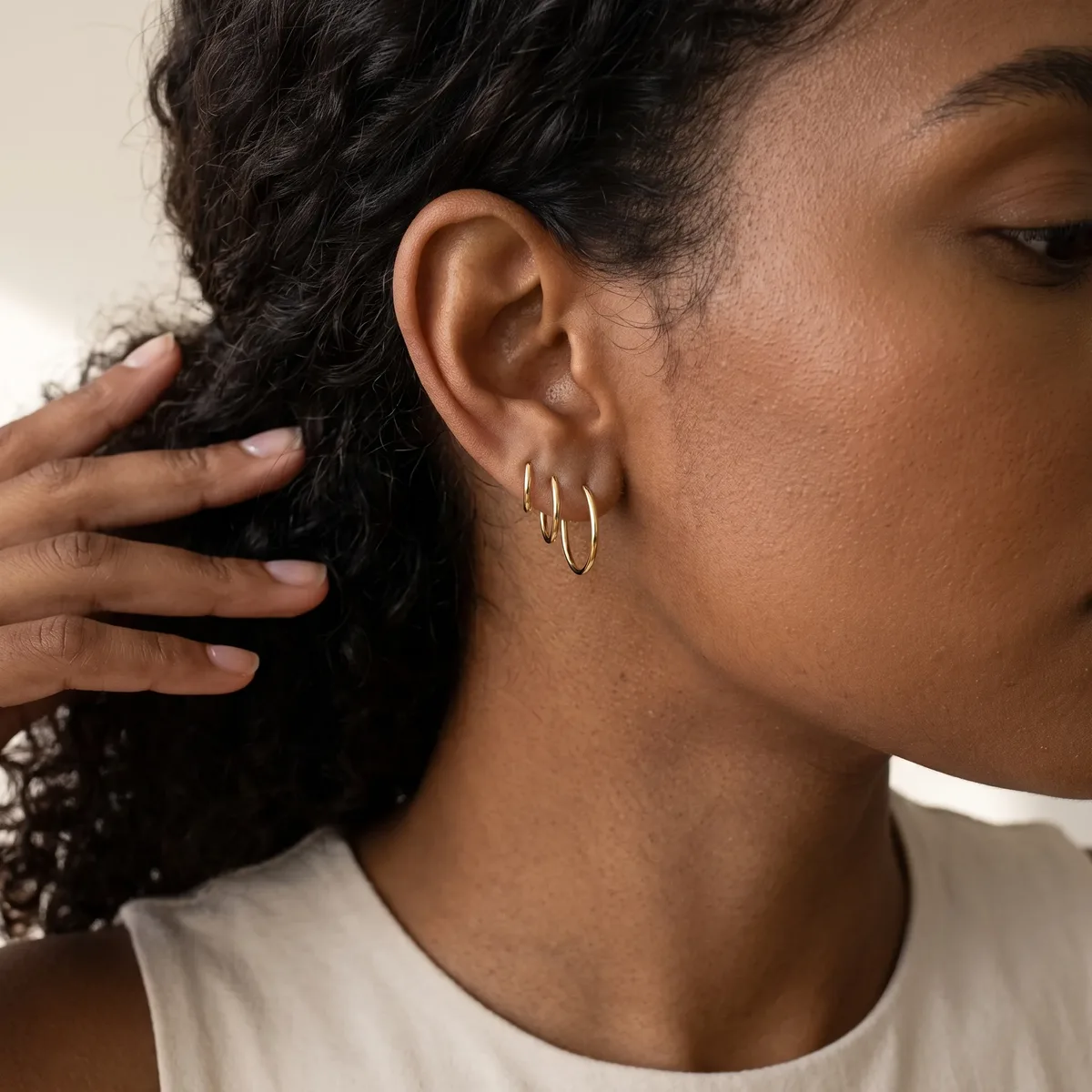 A person with dark curly hair wearing a white t-shirt displays gold hoop earrings on their earlobe while touching their hair with one hand.