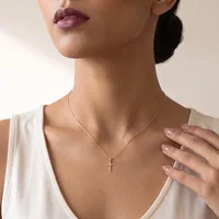 A woman wearing a cream-colored v-neck top displays a gold cross pendant necklace and manicured nails with a neutral polish.