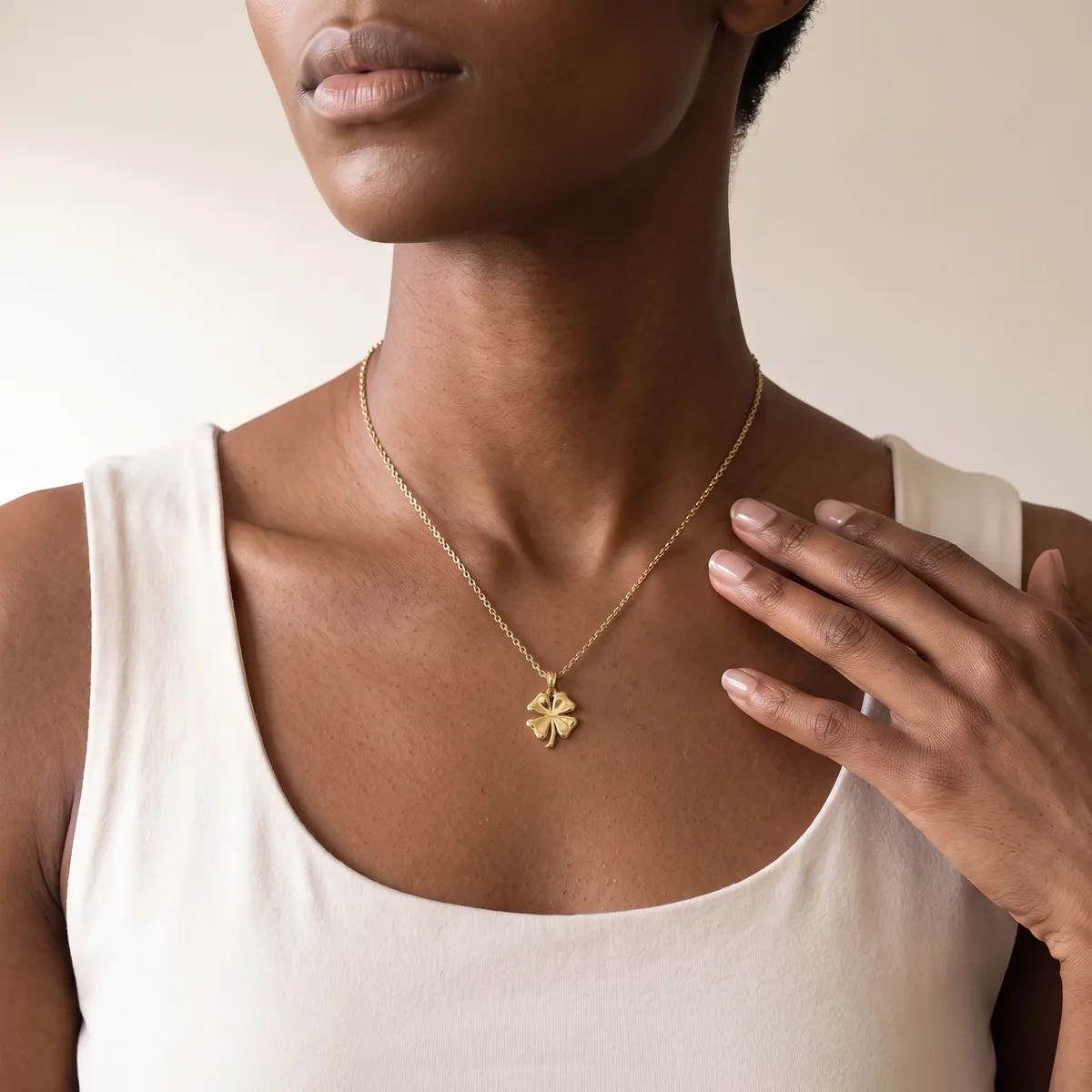 A person wearing a white sleeveless tank top and a gold four-leaf clover pendant necklace, shown from the neck down.