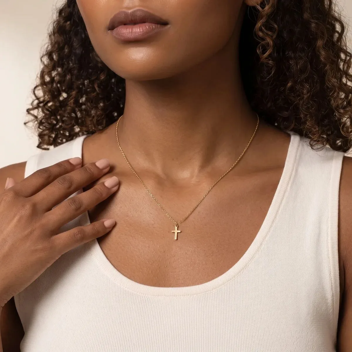 A person wearing a white ribbed tank top and a delicate chain necklace with a small cross pendant rests their hand on their chest.