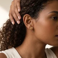 A person with curly dark hair wearing a white sleeveless top displays a small gold hoop earring while touching their hair with one hand.