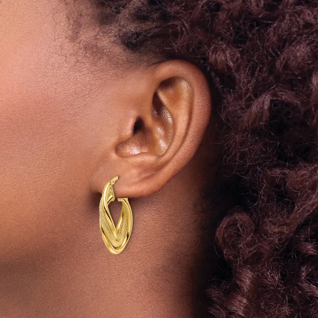 A person wearing a pair of gold-toned hoop earrings with a textured, hammered finish, shown in profile against their ear and dark curled hair.