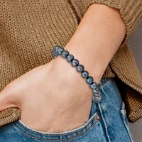 A person wearing a tan knit sweater and blue jeans displays a beaded bracelet made of dark gray pearl-like spheres connected by knotted cord on their wrist.