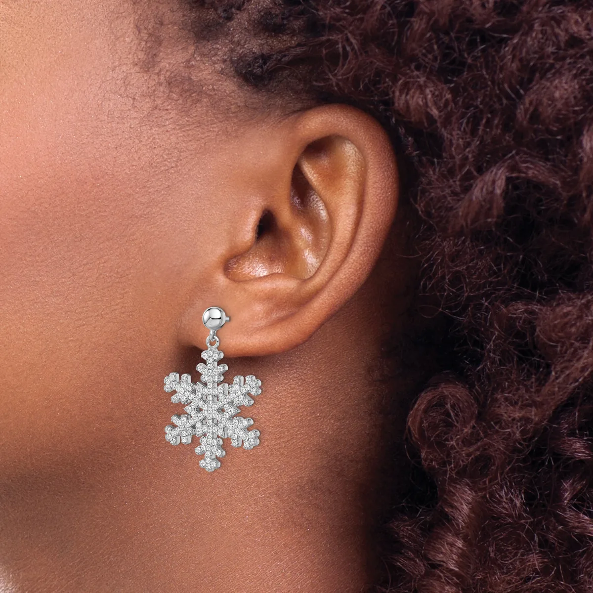 A person wearing a snowflake-shaped drop earring made of rhinestones hanging from a stud post, visible on their earlobe against skin and dark curly hair.