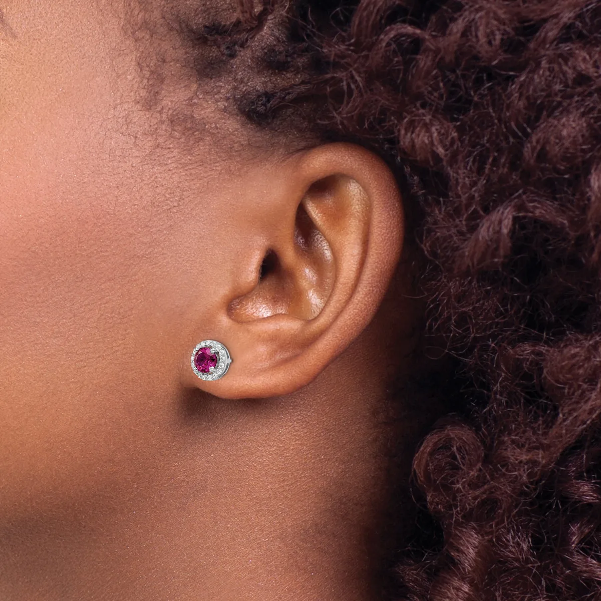 A close-up side view of a person's ear wearing a round stud earring with a red gemstone center surrounded by small white stones in a silver-toned metal setting.