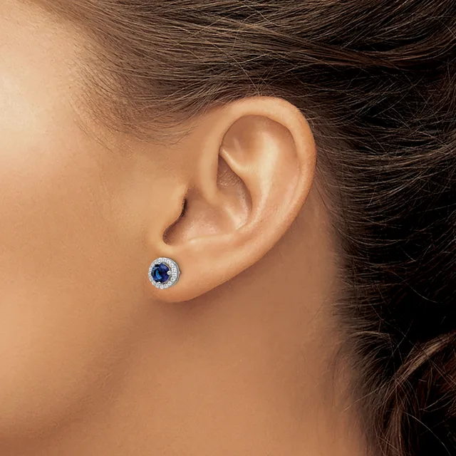 A person wearing a small round stud earring with a dark blue stone surrounded by a halo of smaller diamonds in white metal, shown on their earlobe.