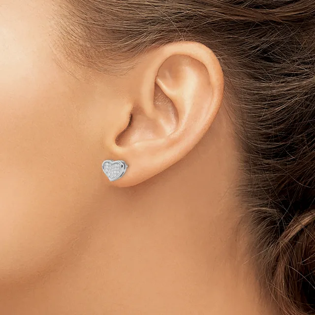 A small heart-shaped stud earring with a textured surface in silver-tone metal is worn in the earlobe of a person with brown hair.