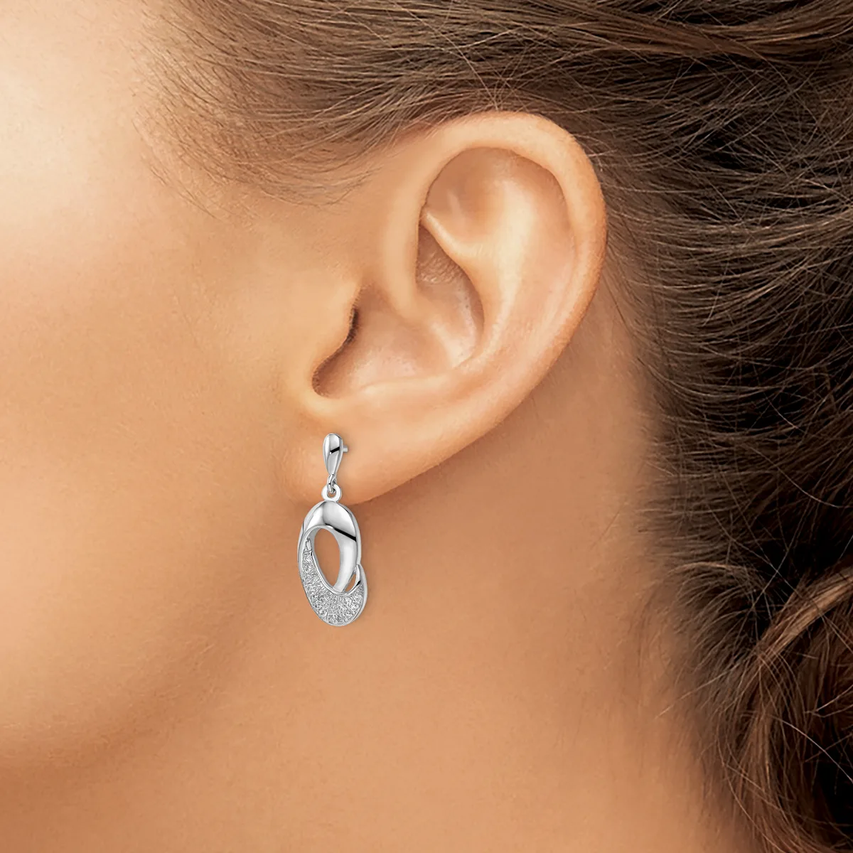 A person wearing a silver drop earring with interlocking oval loops and textured surface detail, shown from the side view of their ear.
