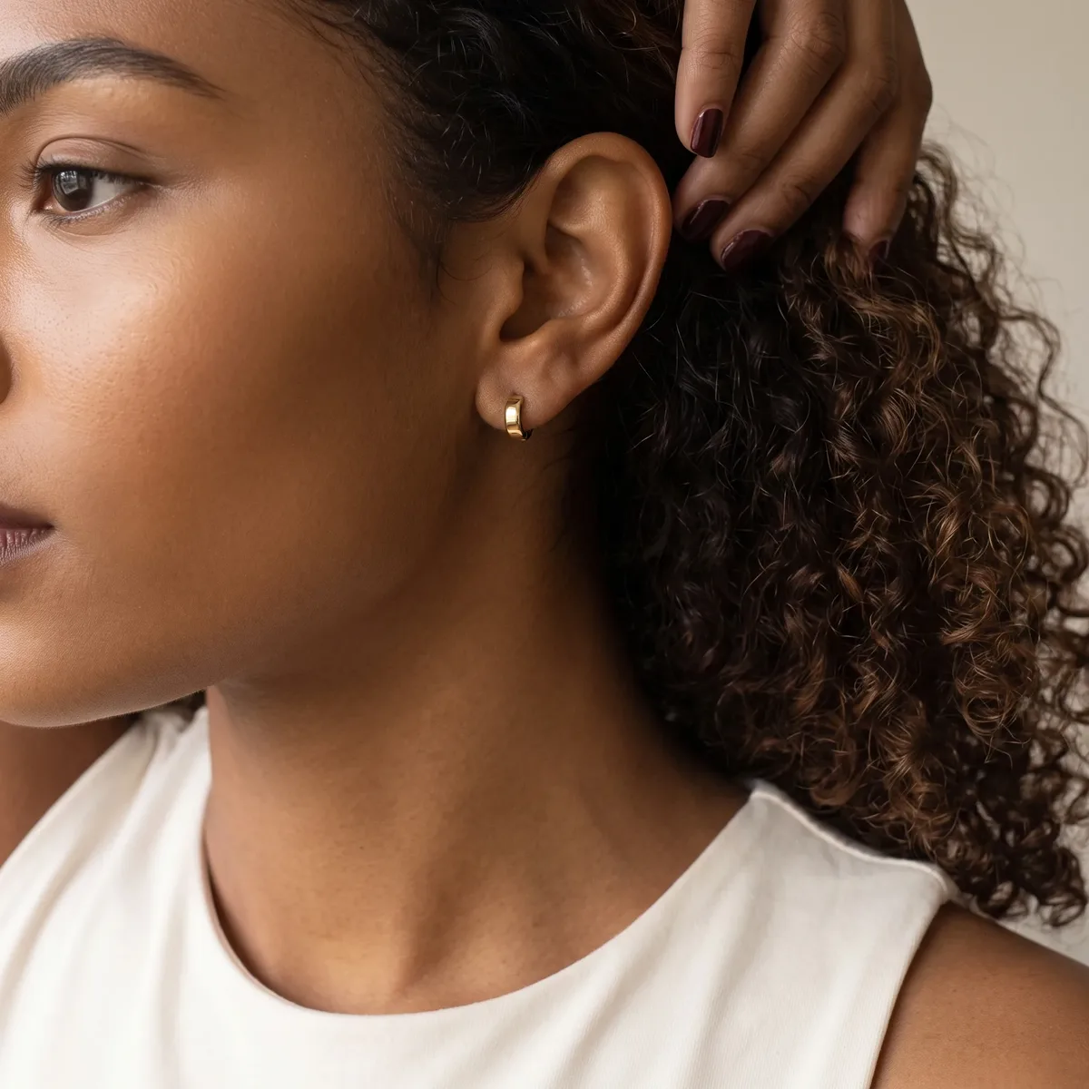 A woman with curly dark hair wearing a white sleeveless top displays a small gold hoop earring in her pierced ear.
