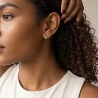A woman with curly dark hair wearing a white sleeveless top displays a small gold hoop earring in her pierced ear.