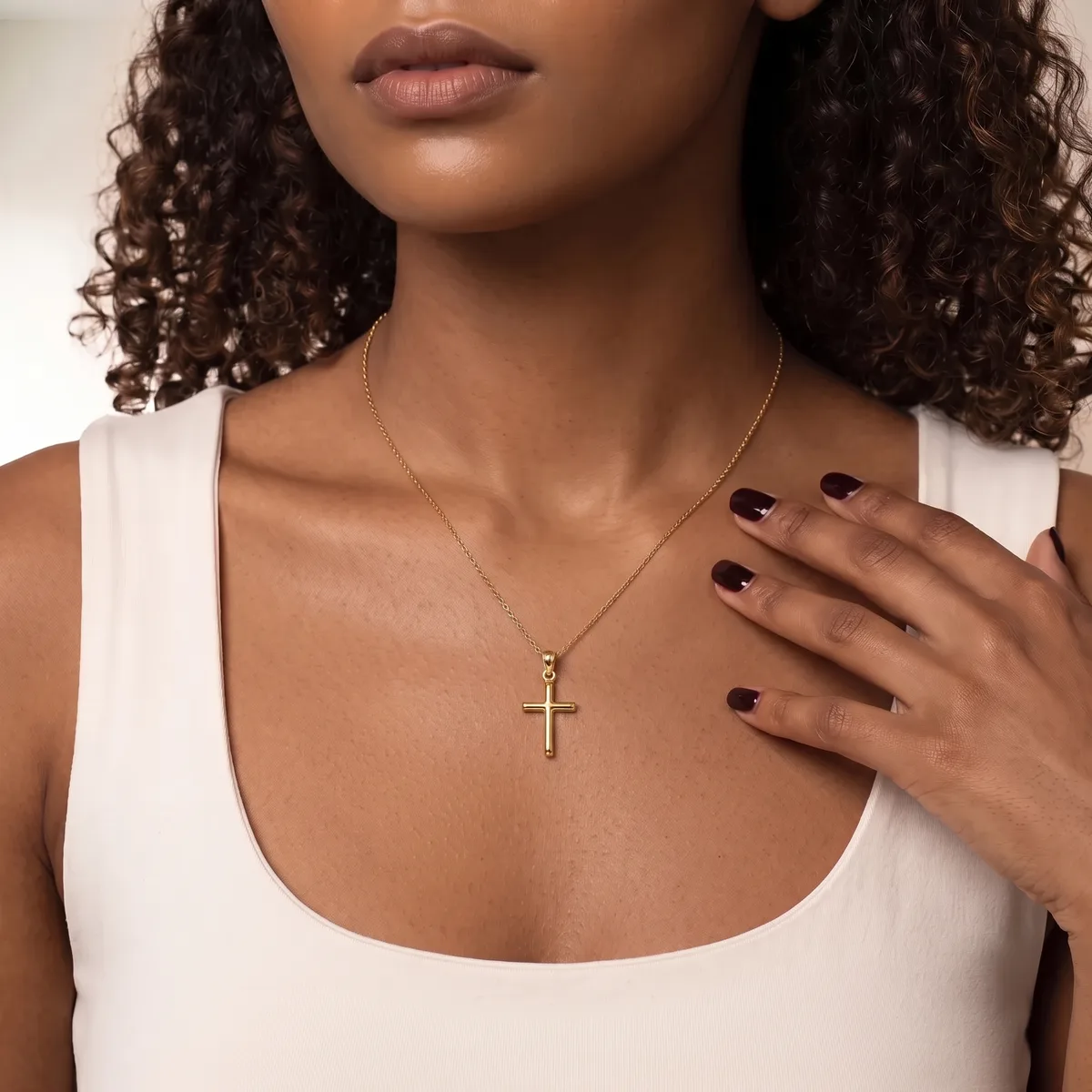 Woman wearing a white sleeveless top and a silver cross necklace, with dark curled hair and dark nail polish, shown from shoulders up against a neutral background.