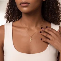 Woman wearing a white sleeveless top and a silver cross necklace, with dark curled hair and dark nail polish, shown from shoulders up against a neutral background.