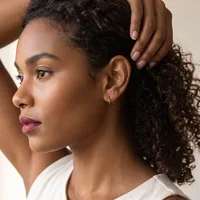 A person with curly dark hair pulled back with one hand, wearing a white top, shown in profile against a neutral background.