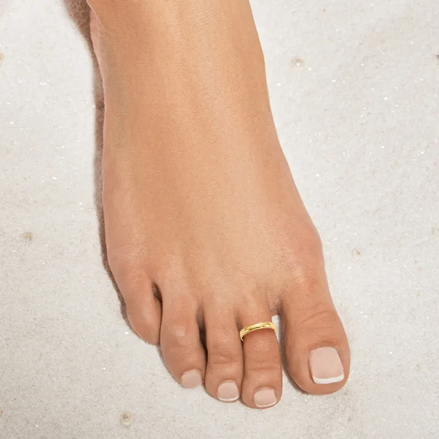 A woman's foot on sand wearing a gold band ring on her fourth toe with a natural nail polish manicure.