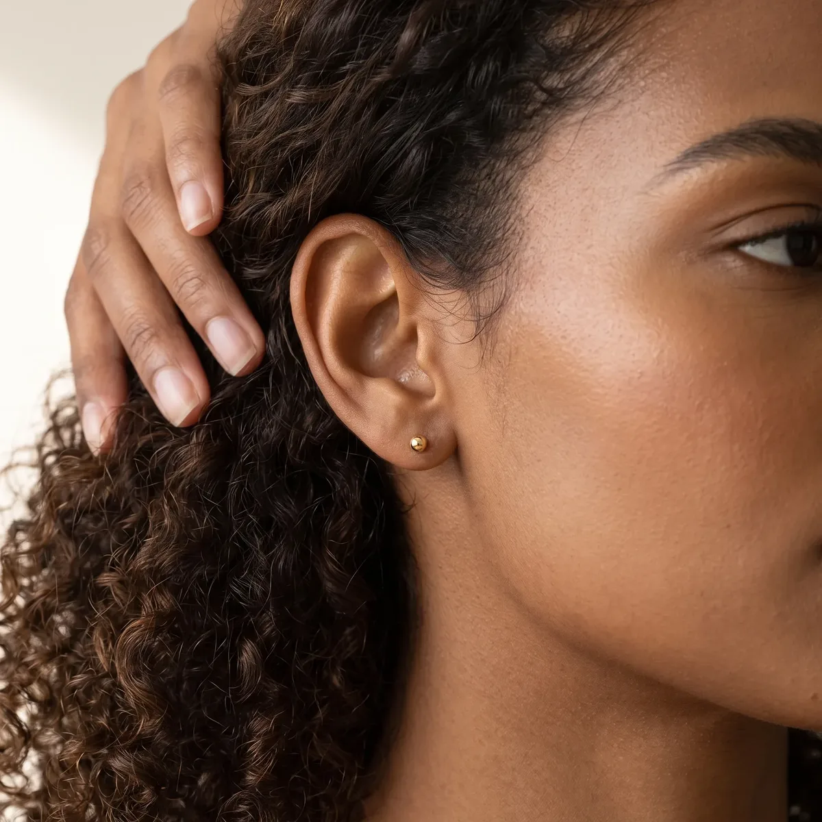 A person with curly dark hair and medium skin tone wears a small round stud earring in their earlobe while touching their hair with their hand.