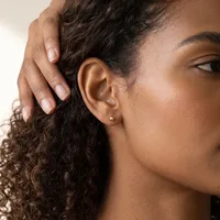 A person with curly dark hair and medium skin tone wears a small round stud earring in their earlobe while touching their hair with their hand.