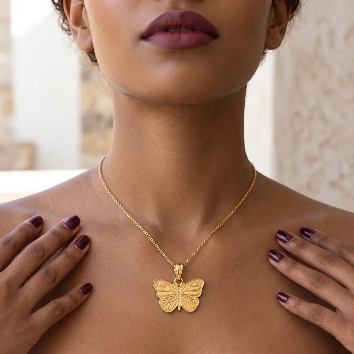 A woman wearing a gold butterfly pendant necklace on a delicate chain, with dark red lipstick and matching nail polish, hands resting on her chest.
