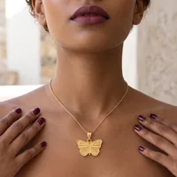 A woman wearing a gold butterfly pendant necklace on a delicate chain, with dark red lipstick and matching nail polish, hands resting on her chest.