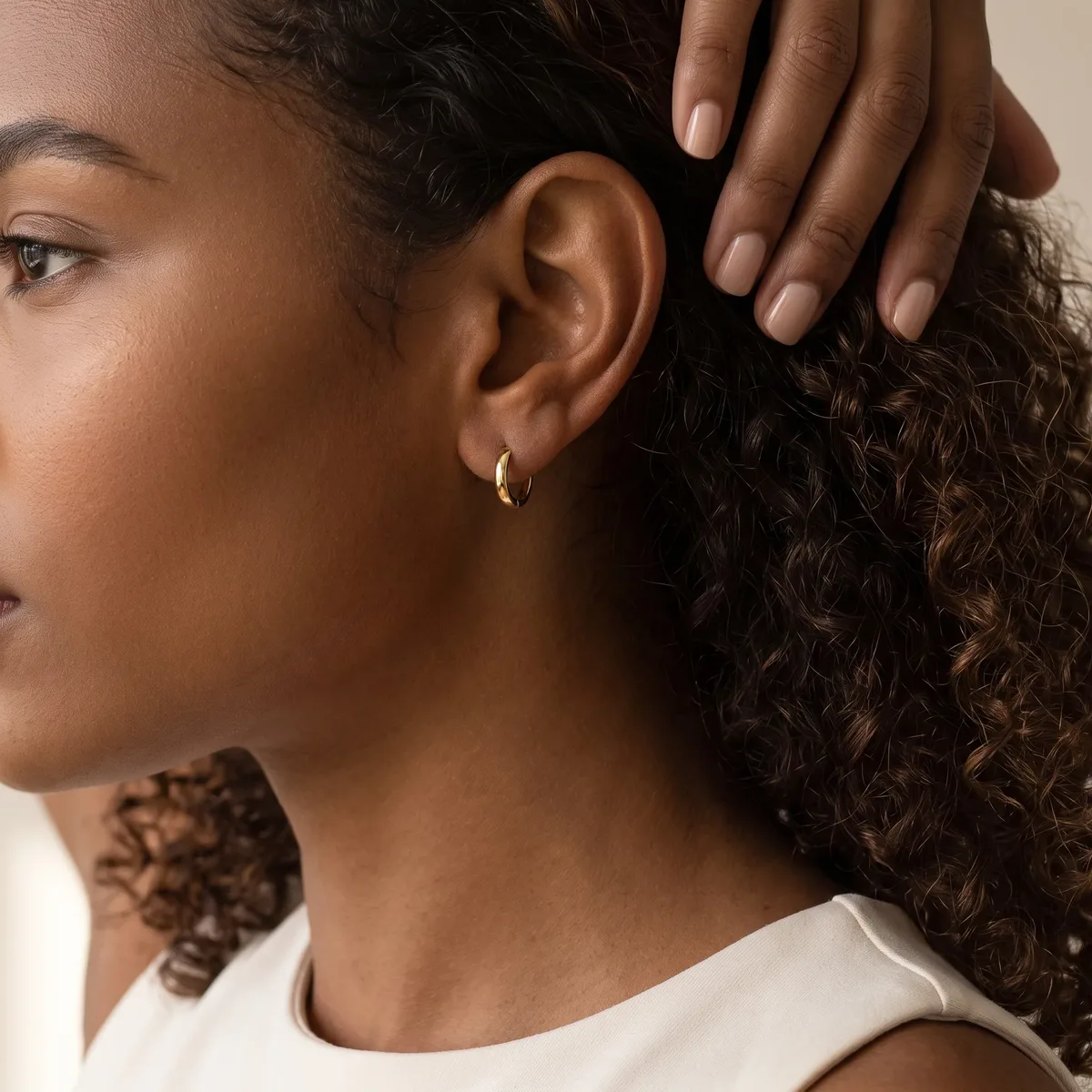 A person with curly brown hair wearing a white sleeveless top and small gold hoop earrings, shown in profile from the side of their face and neck.