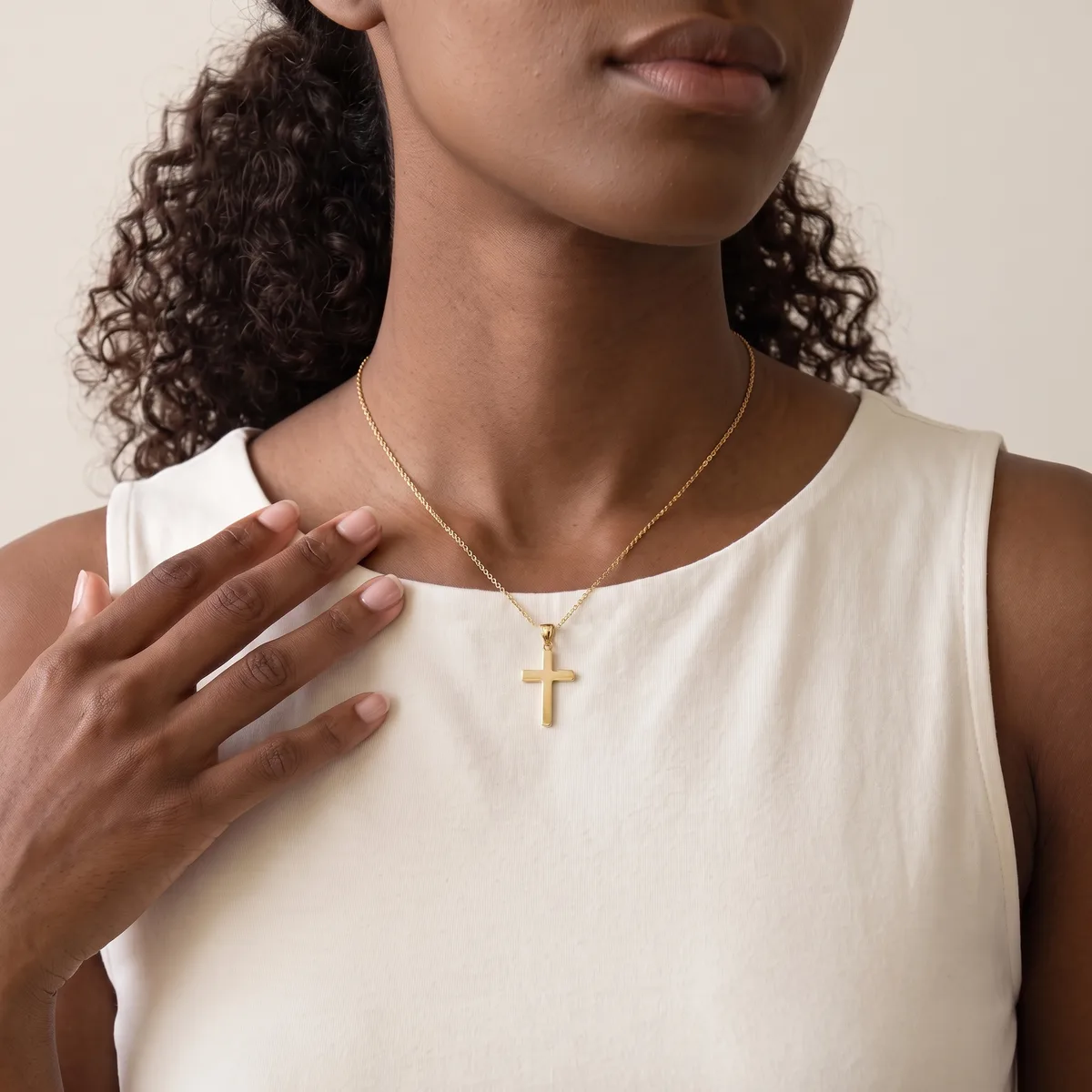 A woman with long curly dark hair wearing a cream sleeveless tank top and a gold cross pendant necklace on a thin chain.