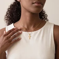 A woman with long curly dark hair wearing a cream sleeveless tank top and a gold cross pendant necklace on a thin chain.