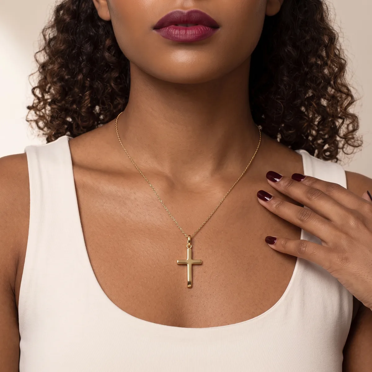 A woman wearing a white sleeveless top and a gold cross pendant necklace, with burgundy lipstick and dark nail polish.