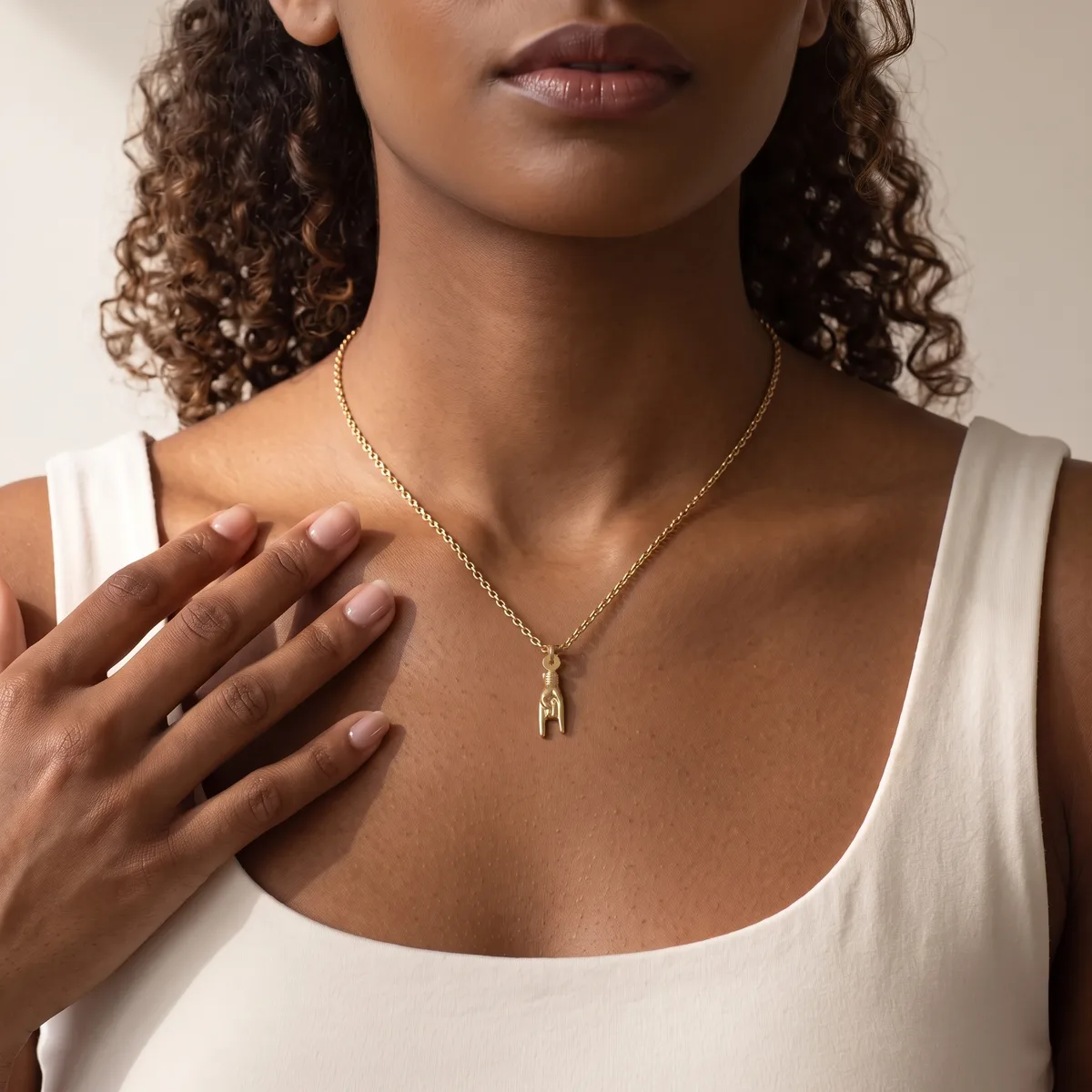 A woman wearing a white sleeveless tank top and a gold chain necklace with a small rectangular pendant, touching her chest with her hand.