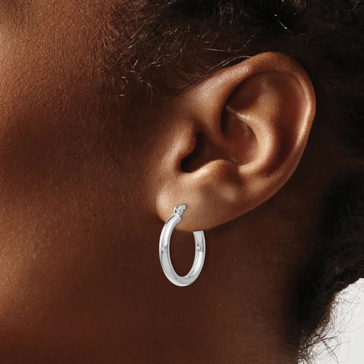 A person wearing a pair of polished silver hoop earrings with a textured surface, shown from the side profile against their ear.