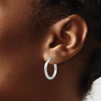 A person wearing a pair of polished silver hoop earrings with a textured surface, shown from the side profile against their ear.