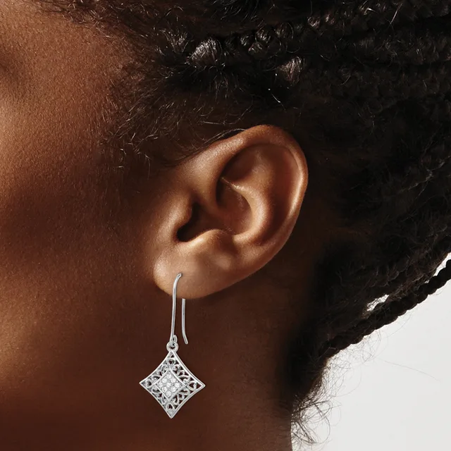 A person wearing a diamond-shaped drop earring with a filigree pattern in silver, suspended from a thin wire hook.