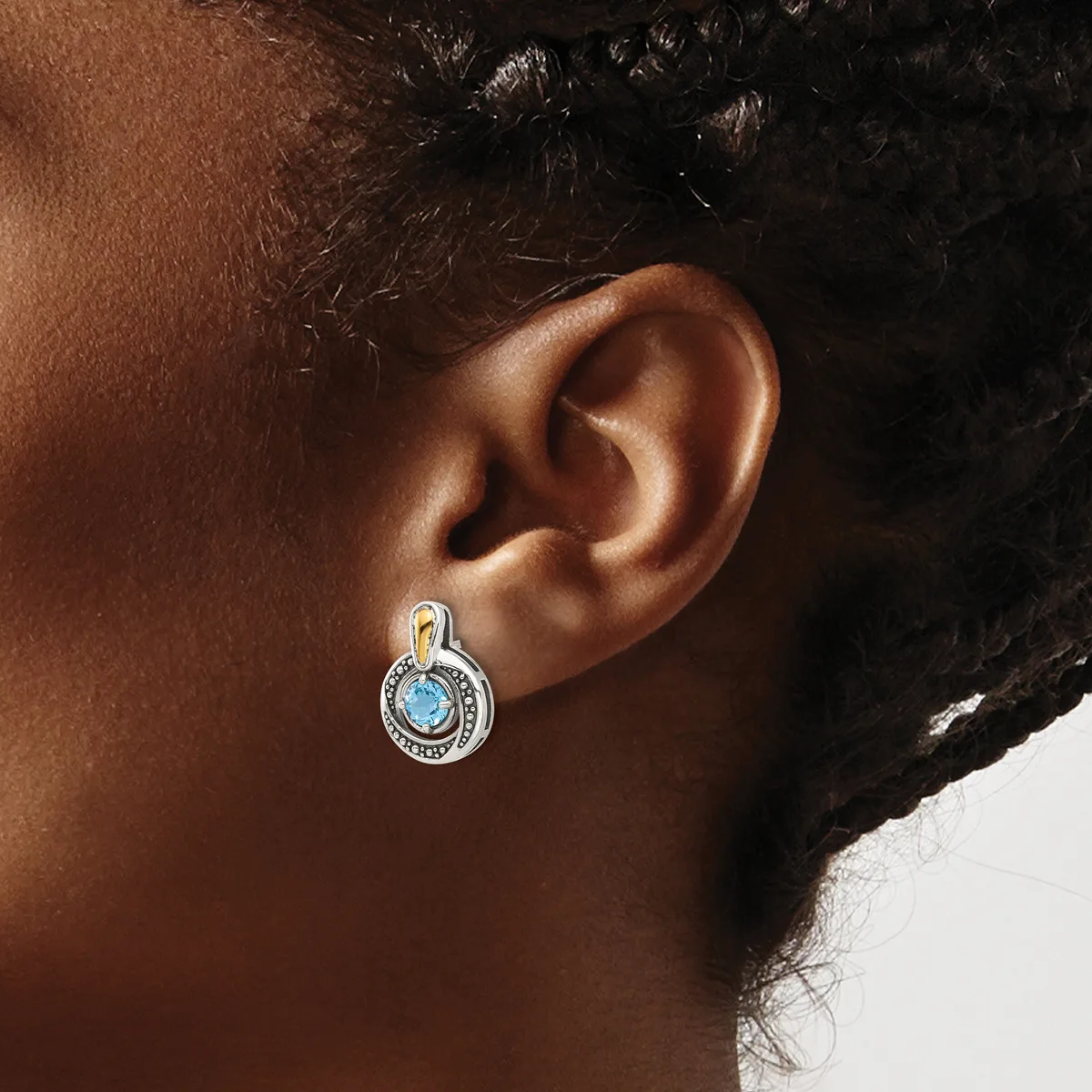 A silver drop earring with a blue circular stone surrounded by small crystals, worn on the ear of a person with brown skin and dark hair.