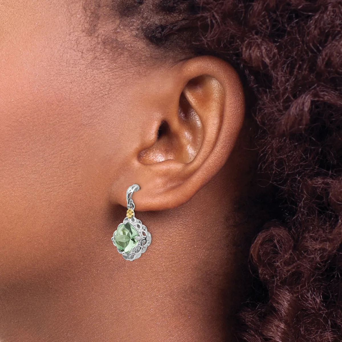 A person wearing a dangling earring with a pale green teardrop-shaped stone set in an ornate silver-toned setting with a curved hook closure.