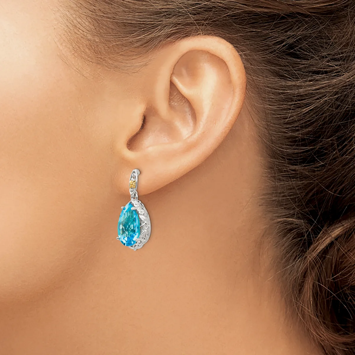 A woman wearing a teardrop-shaped blue topaz drop earring with a diamond-studded silver bail, shown from the side against her ear.