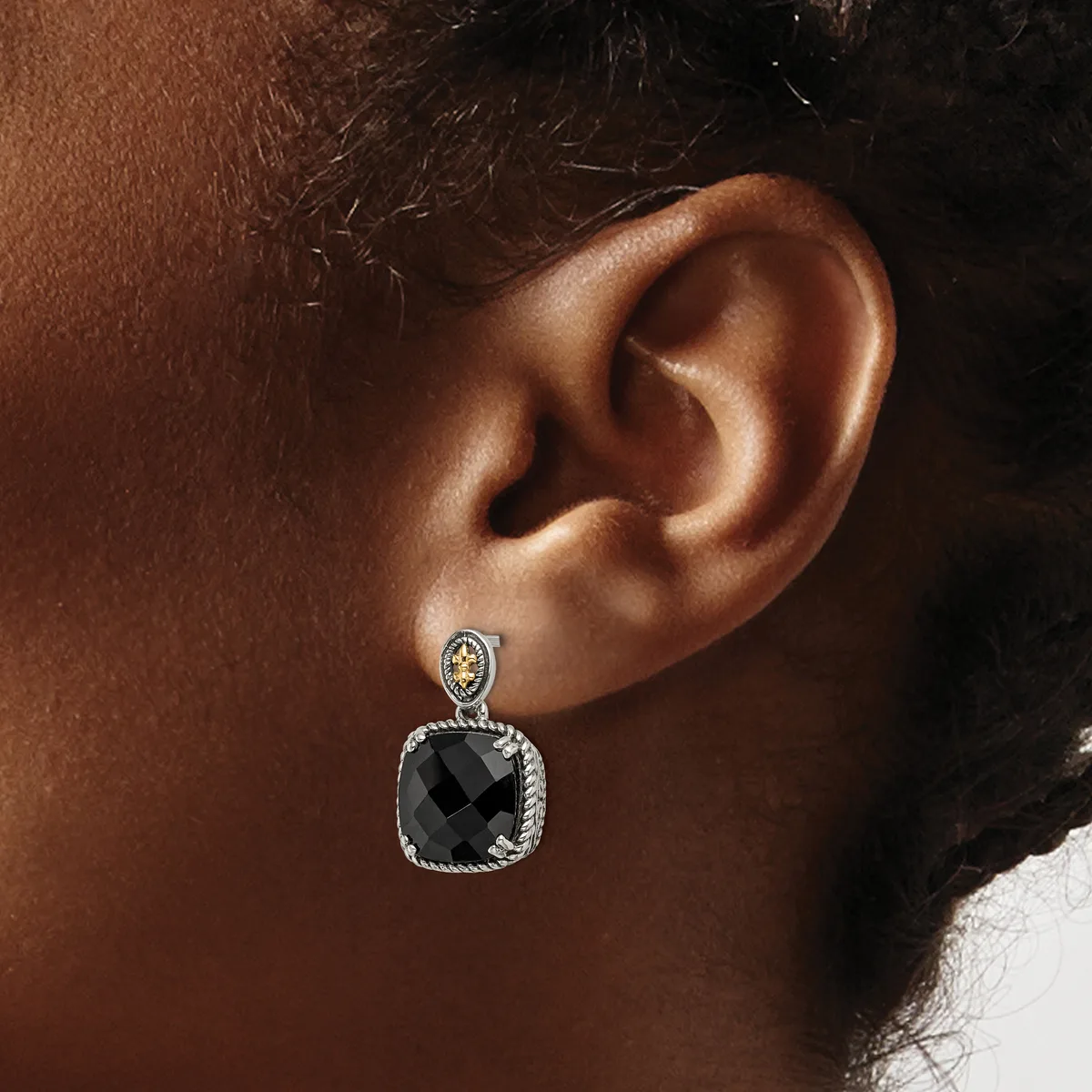 A person wearing a dangling earring with a small round diamond stud at the top and a larger square black faceted stone surrounded by a textured silver bezel below.