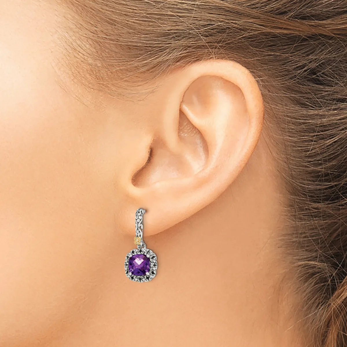 A woman wears a dangling earring with a round purple gemstone surrounded by small diamonds set in white metal, shown on her ear from a side profile view.
