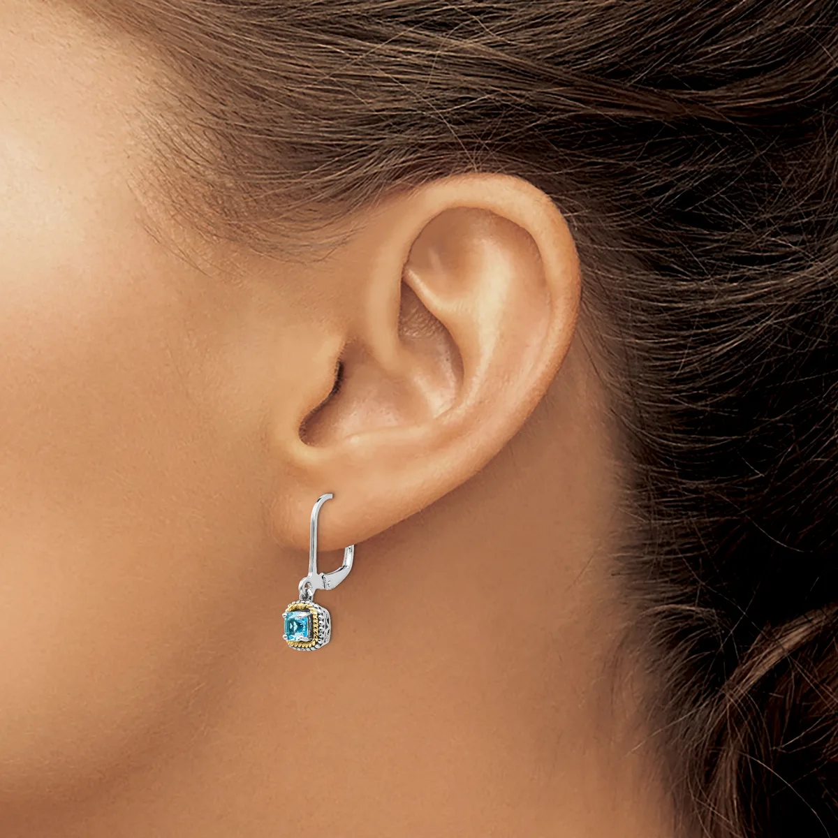 A woman wearing a silver hoop earring with a small square blue stone pendant hanging from it, shown from the side view of her ear and neck.