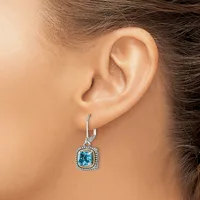 A woman wearing a silver drop earring with a cushion-cut blue topaz stone surrounded by a halo of small diamonds on a lever back hook.