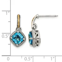 A pair of silver drop earrings with cushion-cut blue topaz stones surrounded by twisted rope detailing and gold accent bands on the posts, shown against a measurement scale.