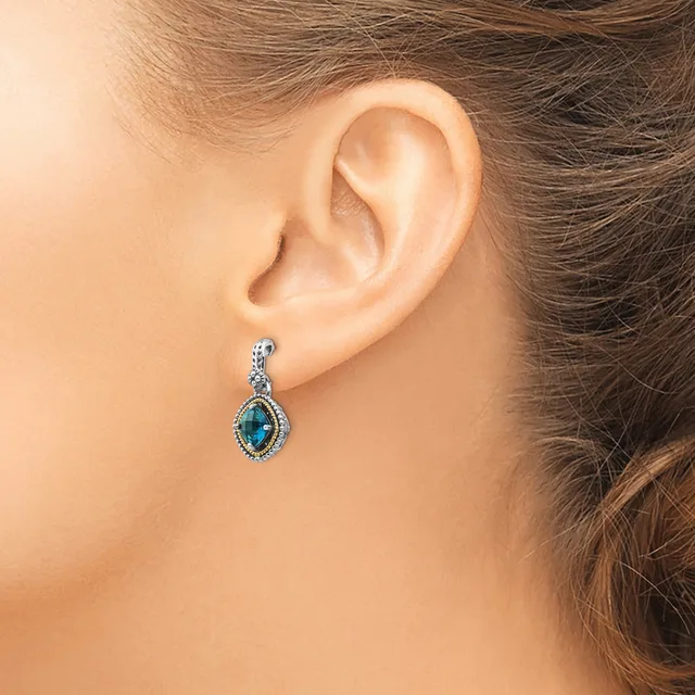 A woman wearing a teardrop-shaped blue gemstone dangle earring with a decorative silver-toned setting and small crystals around the border.