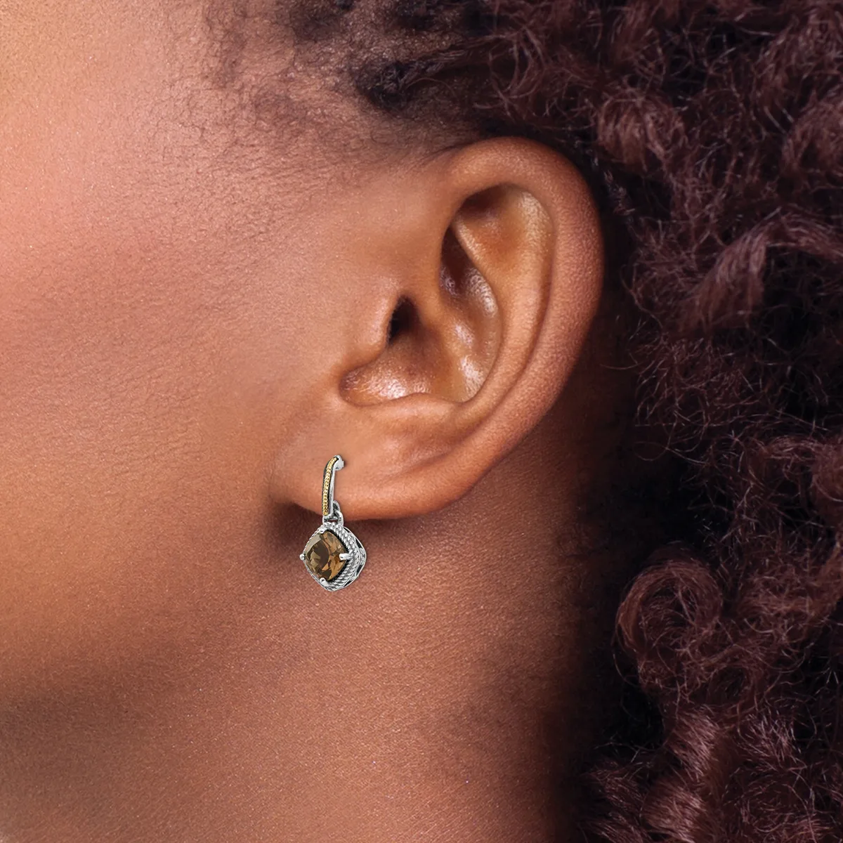 A silver drop earring with a square stone pendant hangs from a person's earlobe, shown in profile against their shoulder and curled dark hair.