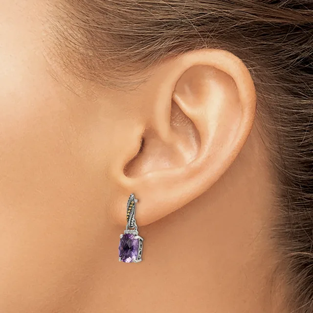 A person wearing a silver drop earring with a purple gemstone and small diamonds on the bail, shown from the side view against their ear.