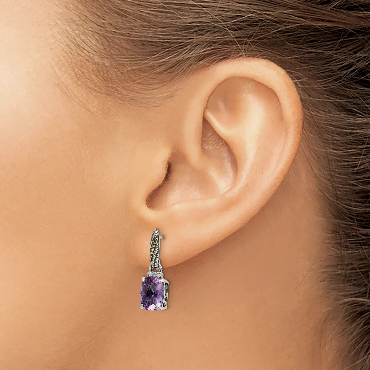 A person wearing a silver drop earring with a purple gemstone and small diamonds on the bail, shown from the side view against their ear.