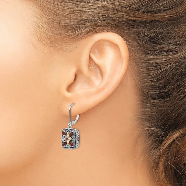A woman wearing a silver drop earring with a square gemstone pendant set in an ornate rectangular frame hangs from a curved hook.